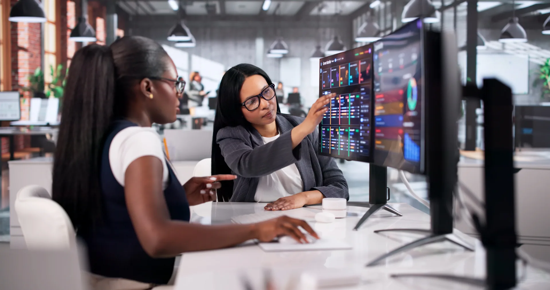Two women professionals collaborate at a desk; reviewing colorful data dashboards on a large monitor in a modern office.