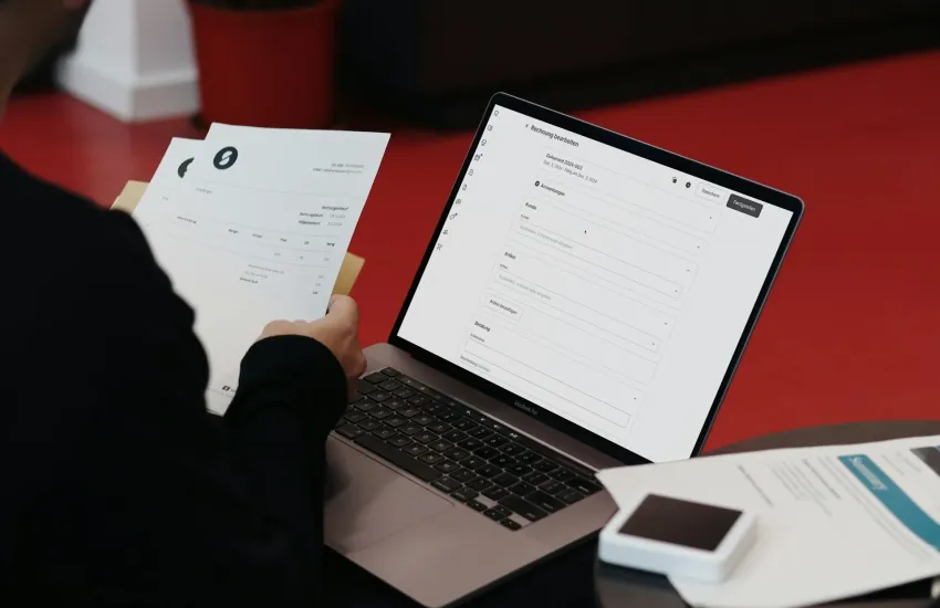 Person holding printed documents beside a laptop that shows a digital document form.