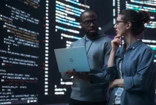 Two colleagues discuss code on a laptop in front of tall screens filled with highlighted source code.